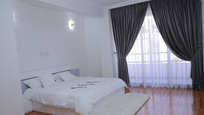 A bedroom with a bed, a white headboard, a wooden floor, and a window with grey curtains. - Abat property (Addis Ababa)