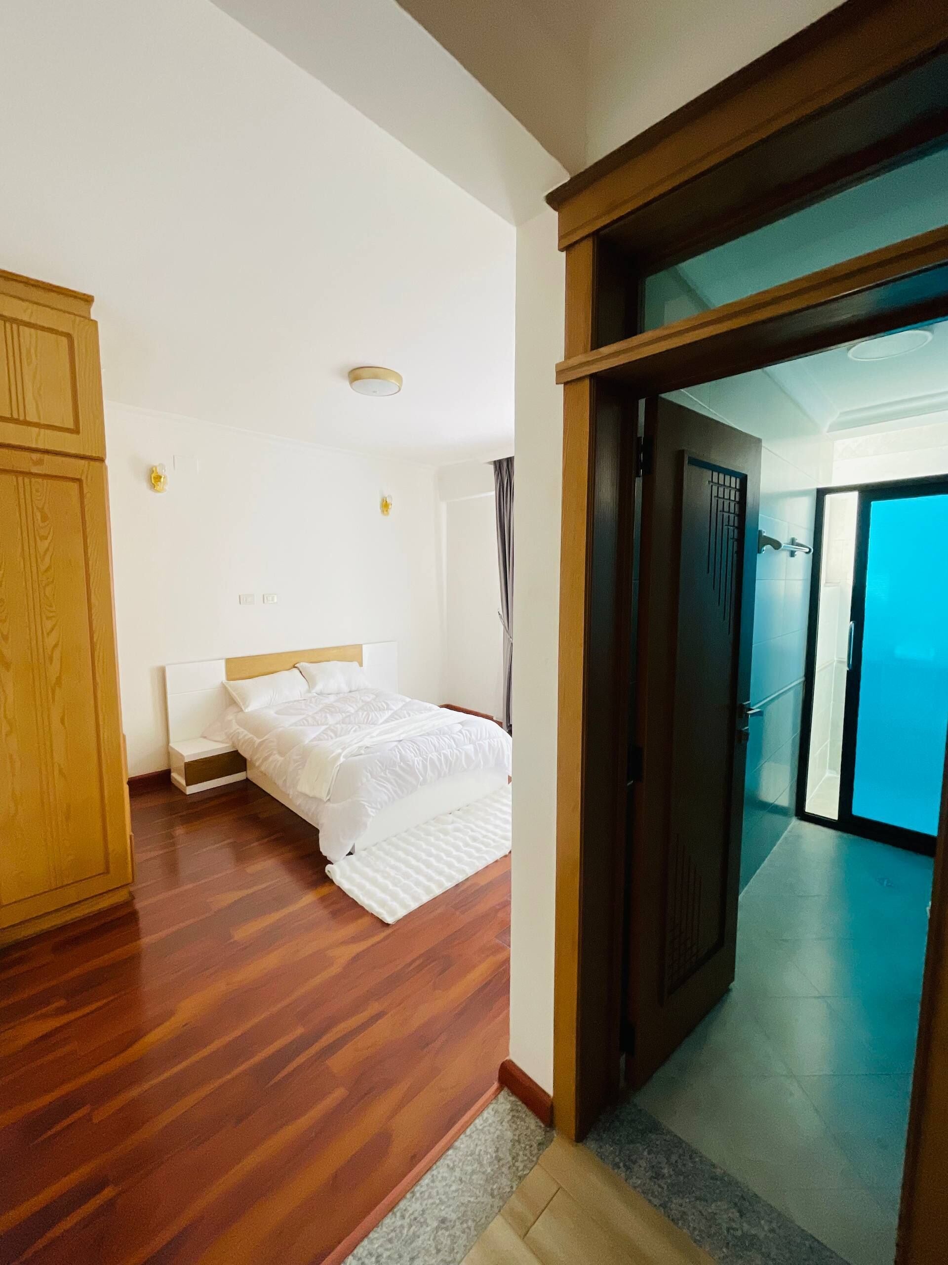 Camera d'albergo con pavimento in legno, un letto con biancheria bianca, un armadio in legno e una porta che dà su un balcone.