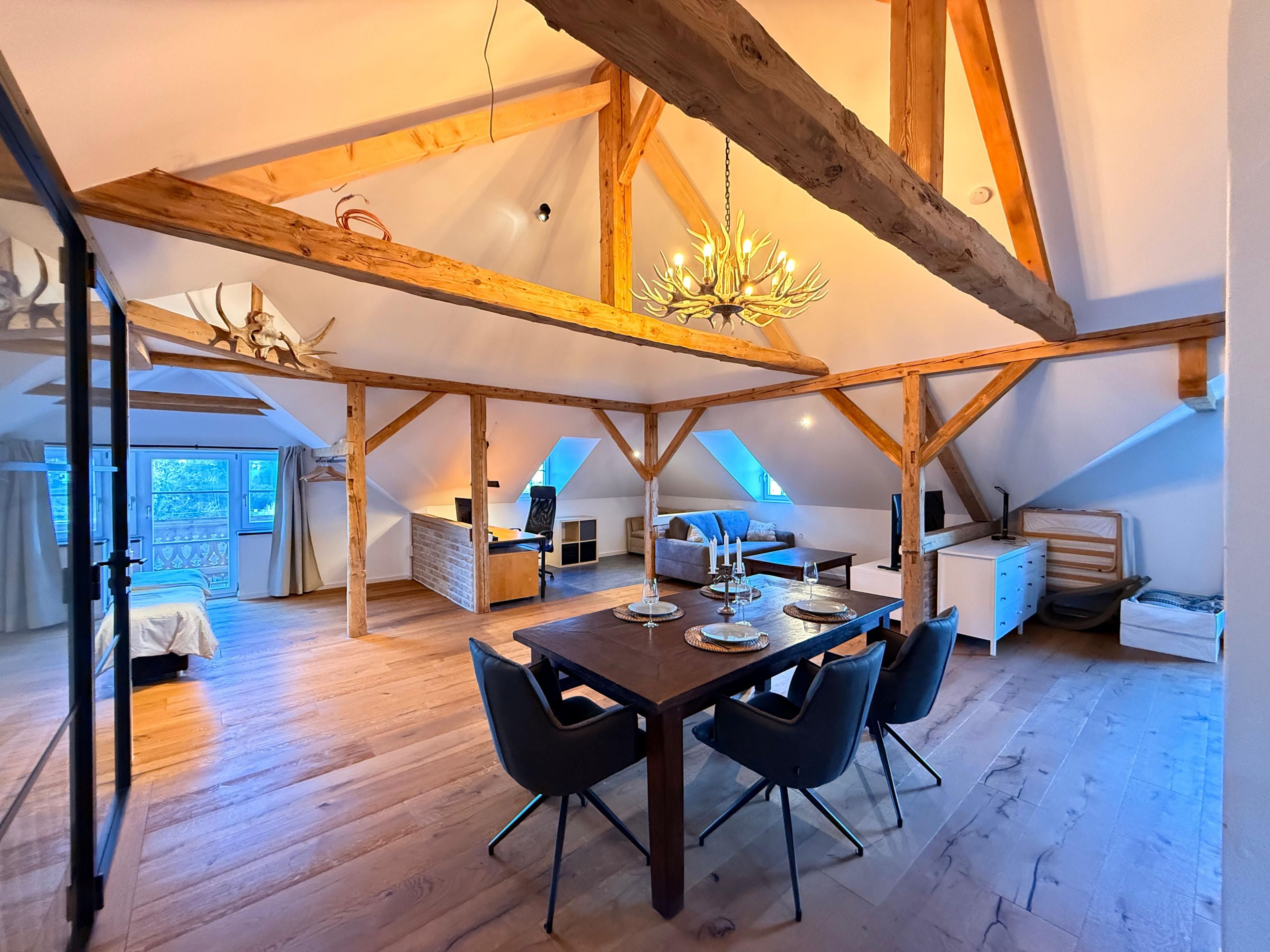 Um loft espaçoso com pé direito alto, vigas de madeira aparentes, uma área de jantar com mesa e cadeiras, e um espaço de estar aconchegante com sofá e lareira.