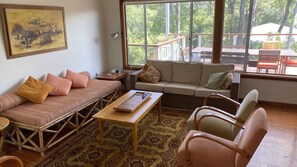 Ein gemütliches Wohnzimmer mit einer Couch, einem kleinen Tisch und einem Sessel. Ein großes Fenster bietet Blick ins Freie.