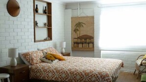 Quarto com colcha estampada, mesa de cabeceira com abajur, estante com objetos decorativos e pintura na parede.
