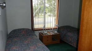 Une chambre avec deux lits, une petite commode en bois et une fenêtre avec des stores.