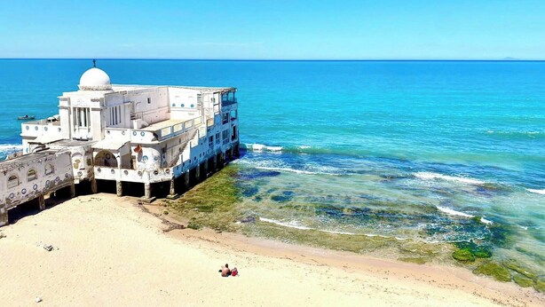 Un bâtiment blanc surmonté d’un dôme, situé sur une plage de sable fin, bordée d’une eau d’un bleu limpide.