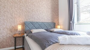 Ein Schlafzimmer mit ornamentalem Wandpapier, einem gepolsterten Kopfteil, zwei Nachttischen mit Lampen und einem Fenster mit Vorhängen.