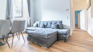 Ein modernes Wohnzimmer mit einem grauen Ecksofa, einem Esstisch mit Stühlen und einem großen Fenster mit Vorhängen.