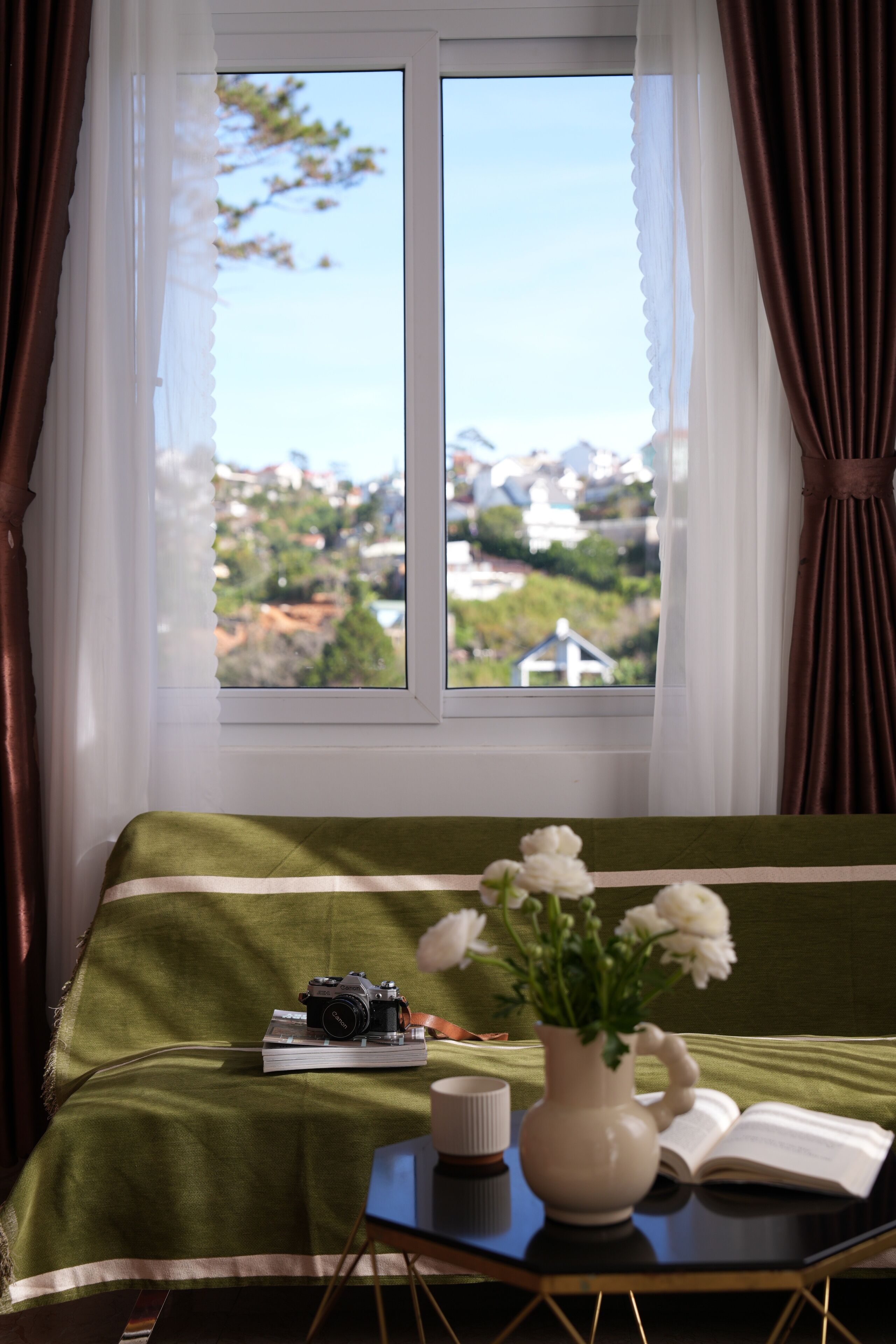 A bed with a green bedspread, a camera, a book, a vase with flowers, and a cup on a side table by a window with curtains.
