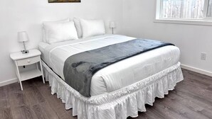A bedroom with a bed, white nightstands, a painting on the wall, and a window with a view of a wooden fence. - Bogachiel Den – Comfortable Forks Getaway (Forks)