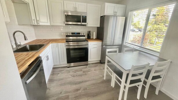 A modern kitchen with white cabinets, stainless steel appliances, a wooden countertop, and a dining area with a table and chairs. - Bogachiel Den – Comfortable Forks Getaway (Forks)