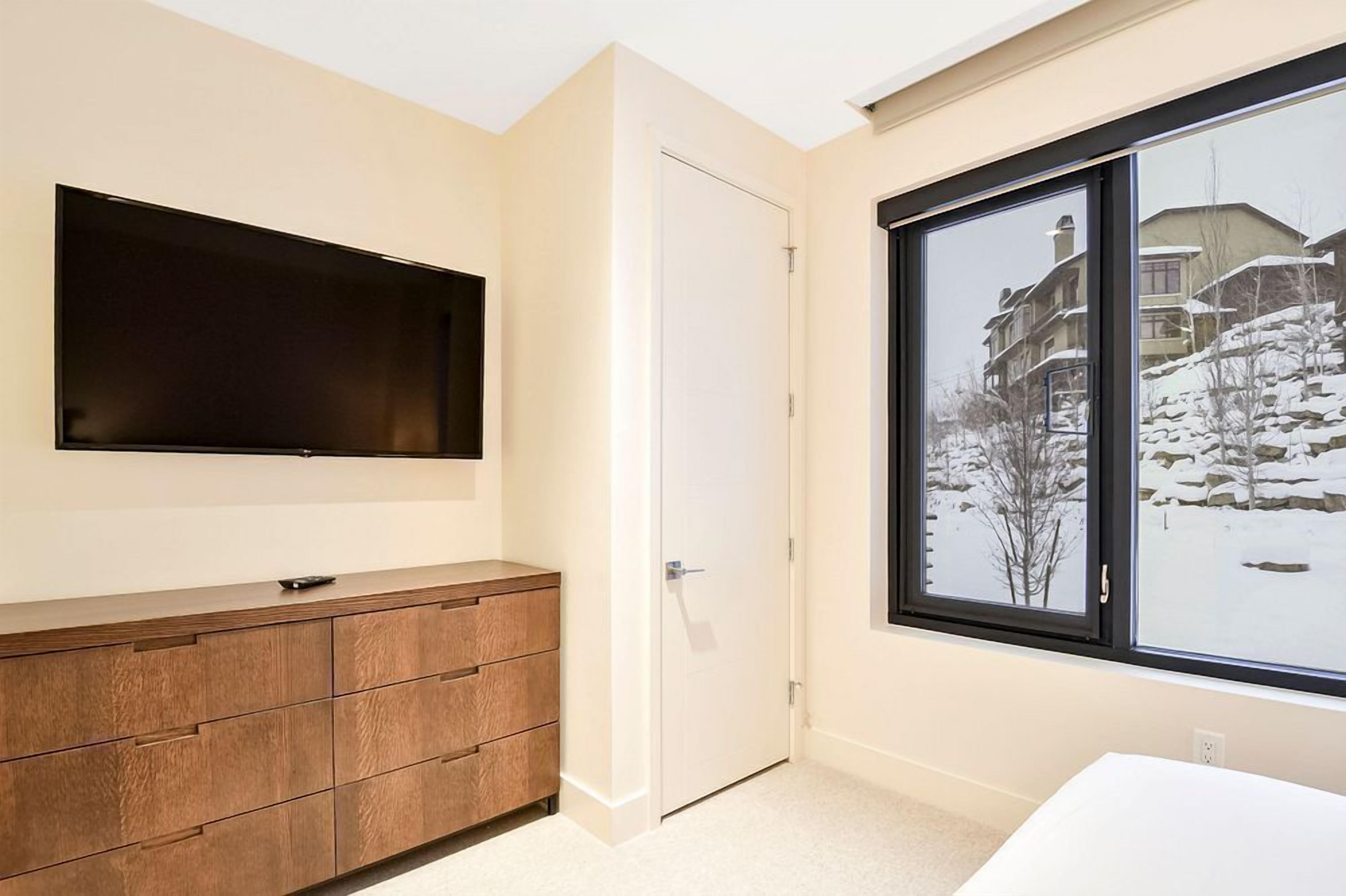 Een slaapkamer met een groot raam, een flatscreen televisie aan de wand en een houten dressoir.