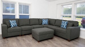 Sofá modular com apoio para os pés combinando e almofadas estampadas, apoiado contra uma parede branca com grandes janelas.