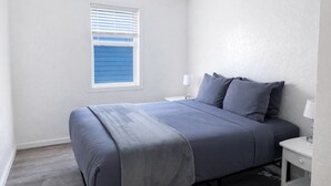 Quarto com cama, mesa de cabeceira, abajur e janela com persianas.
