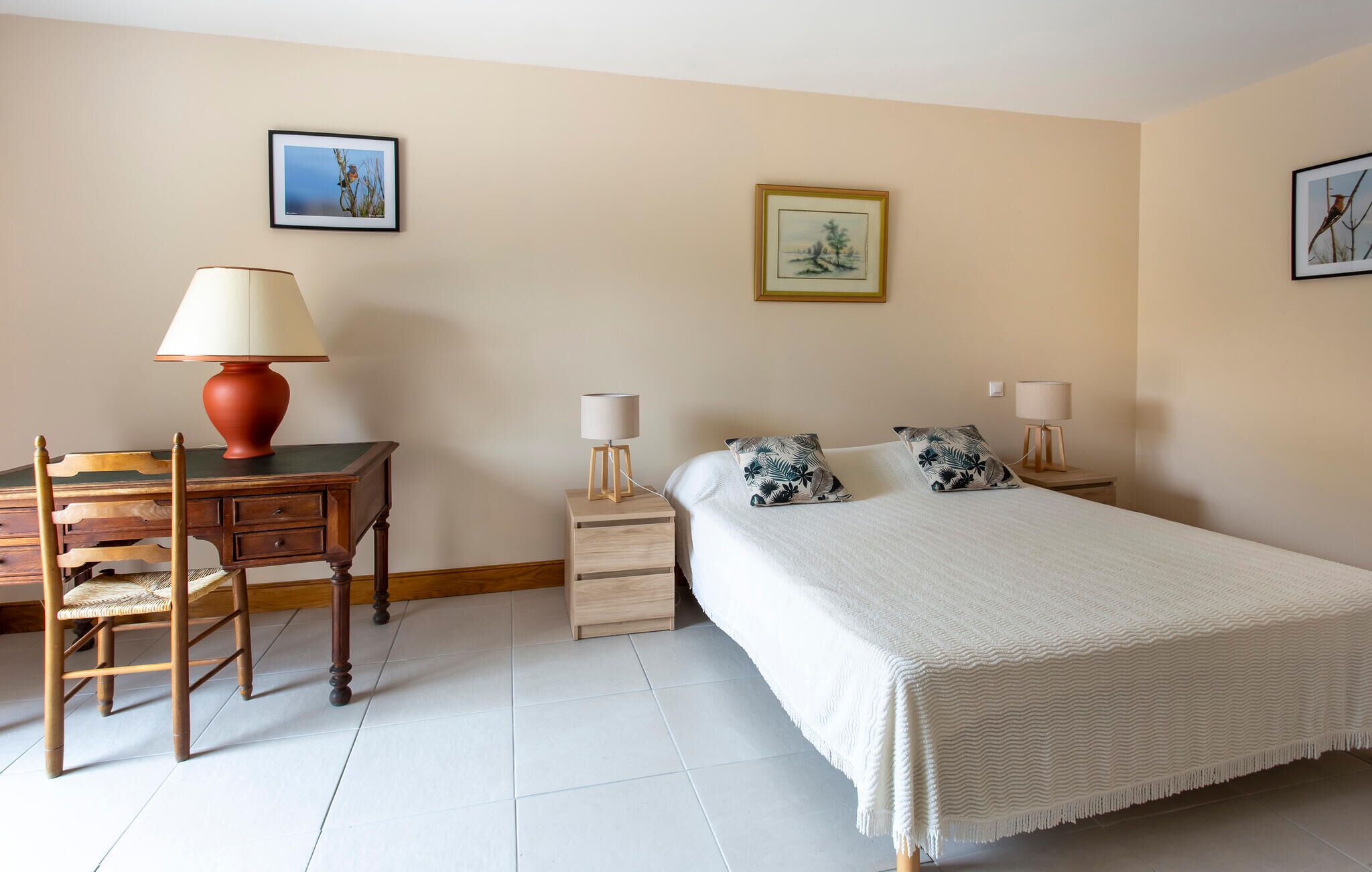 A bedroom with a bed, two bedside tables with lamps, a desk with a chair, and two framed pictures on the wall.