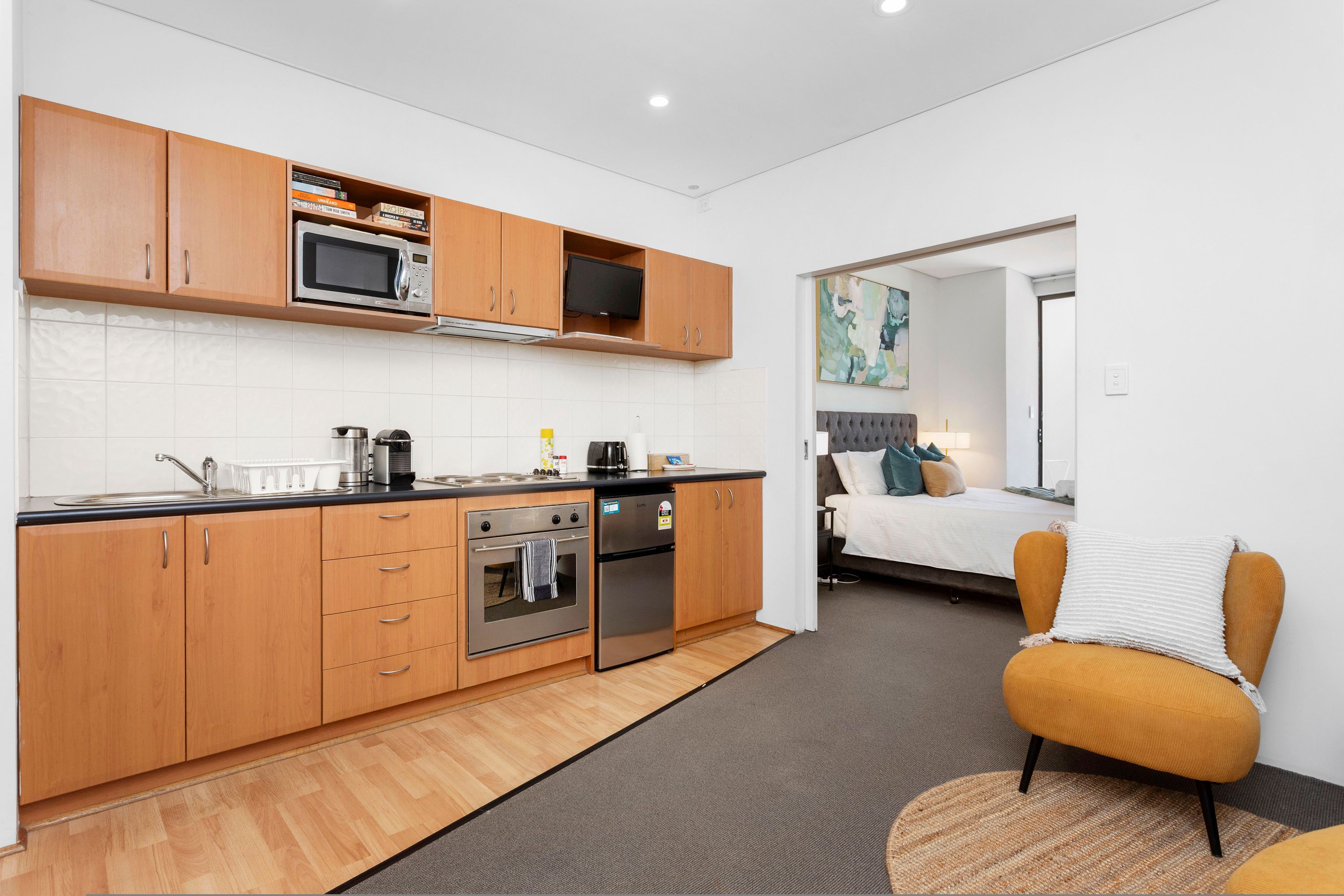 A compact kitchen with wooden cabinets, a microwave, oven, and a refrigerator. A bed with a white pillow and a yellow armchair are visible in the adjacent room.