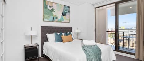 A hotel room with a large bed, two bedside tables, a painting on the wall, and a balcony with a view.
