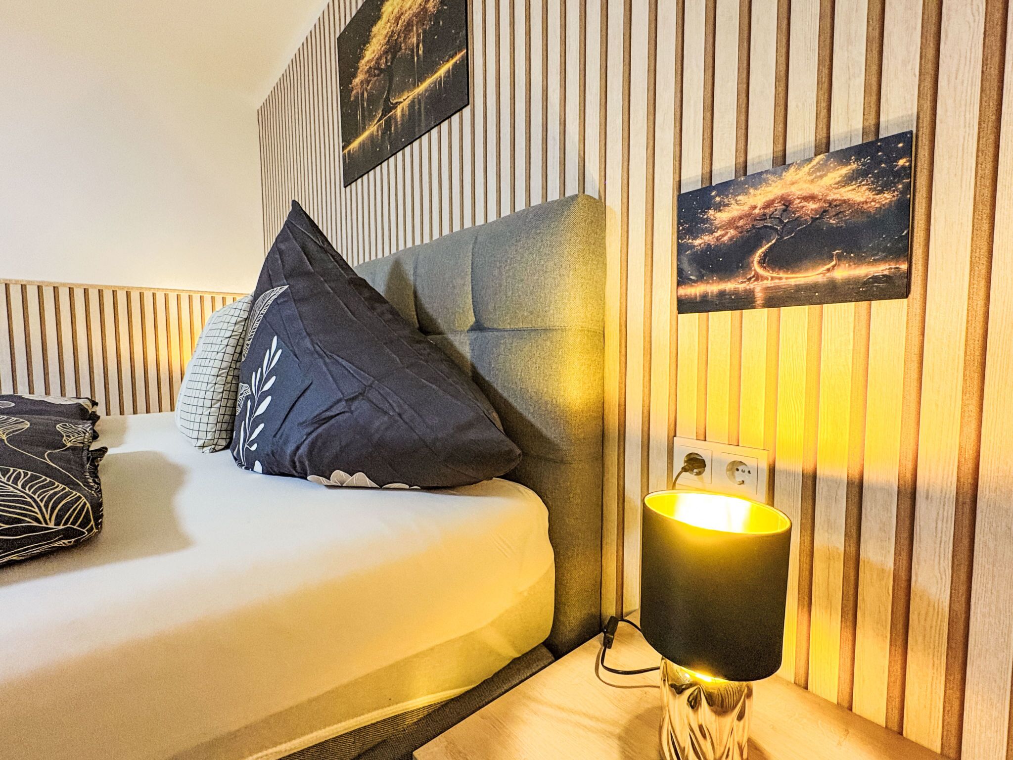 Une chambre à coucher bien rangée, avec un lit, des oreillers, une lampe de chevet et deux œuvres d’art accrochées au mur.