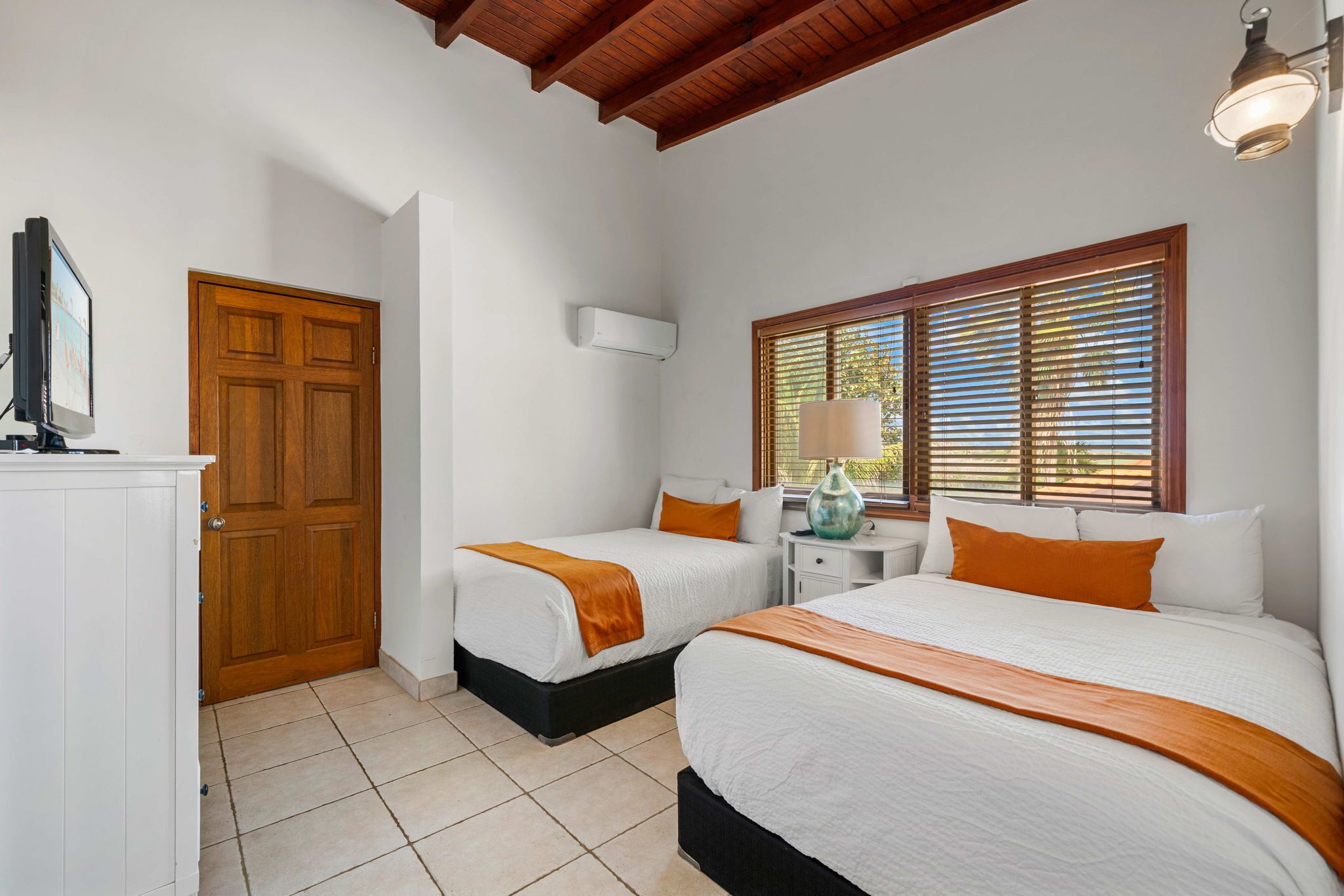 A bedroom with two beds, a wooden door, a white cabinet, and a window with blinds.