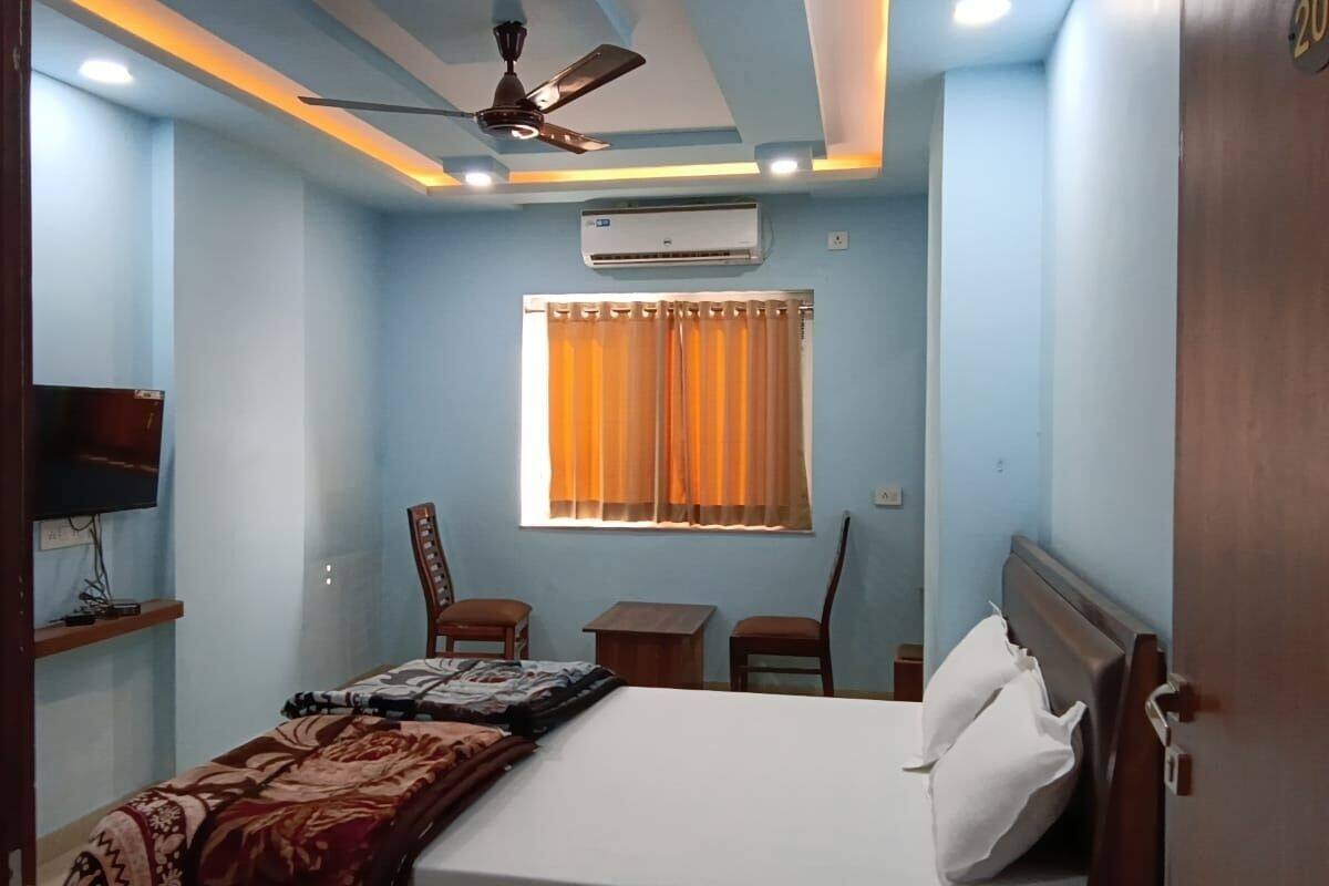 A bedroom with a bed, a desk, two chairs, a ceiling fan, an air conditioner, and a TV on a shelf.