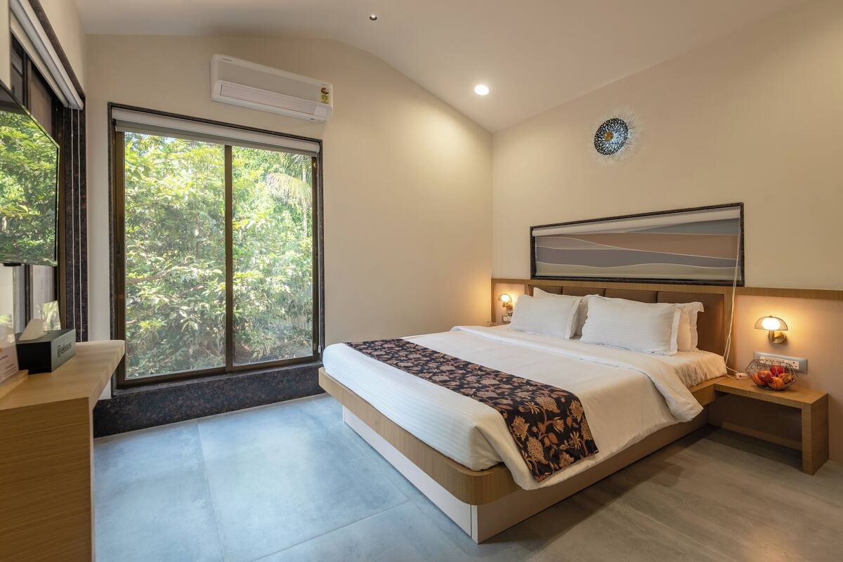 A modern hotel room with a large bed, a bedside table with a fruit bowl, a nightstand with a lamp, and a view of greenery through a large window.