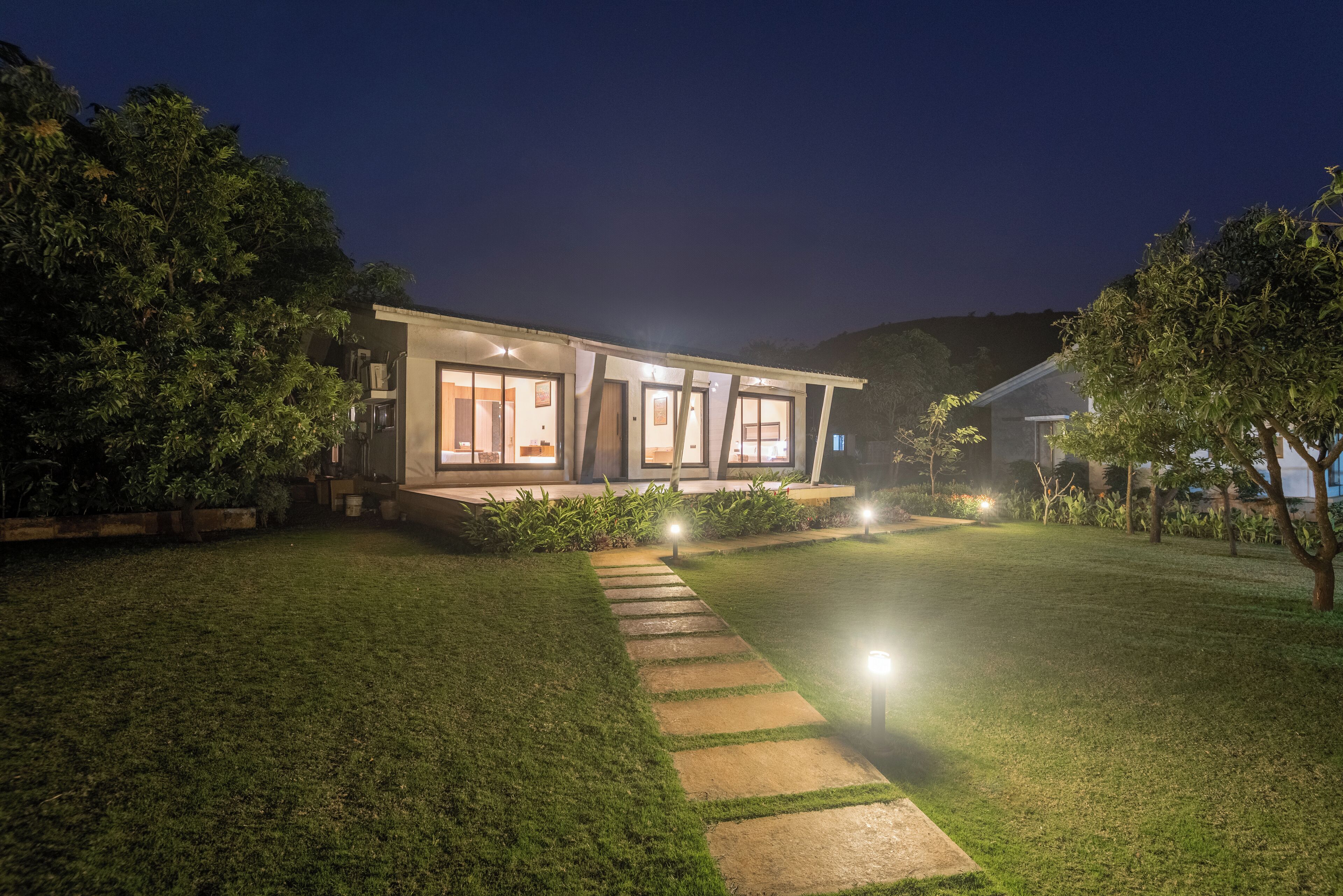 A modern house with a well-lit exterior, a stone pathway leading to the entrance, and a lush green lawn.
