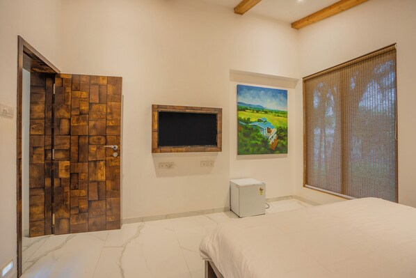 A bedroom with a bed, a small white refrigerator, a wooden door, a painting, and a window with blinds. - ELIVAAS Energise Soul Retreat (Trimbak)