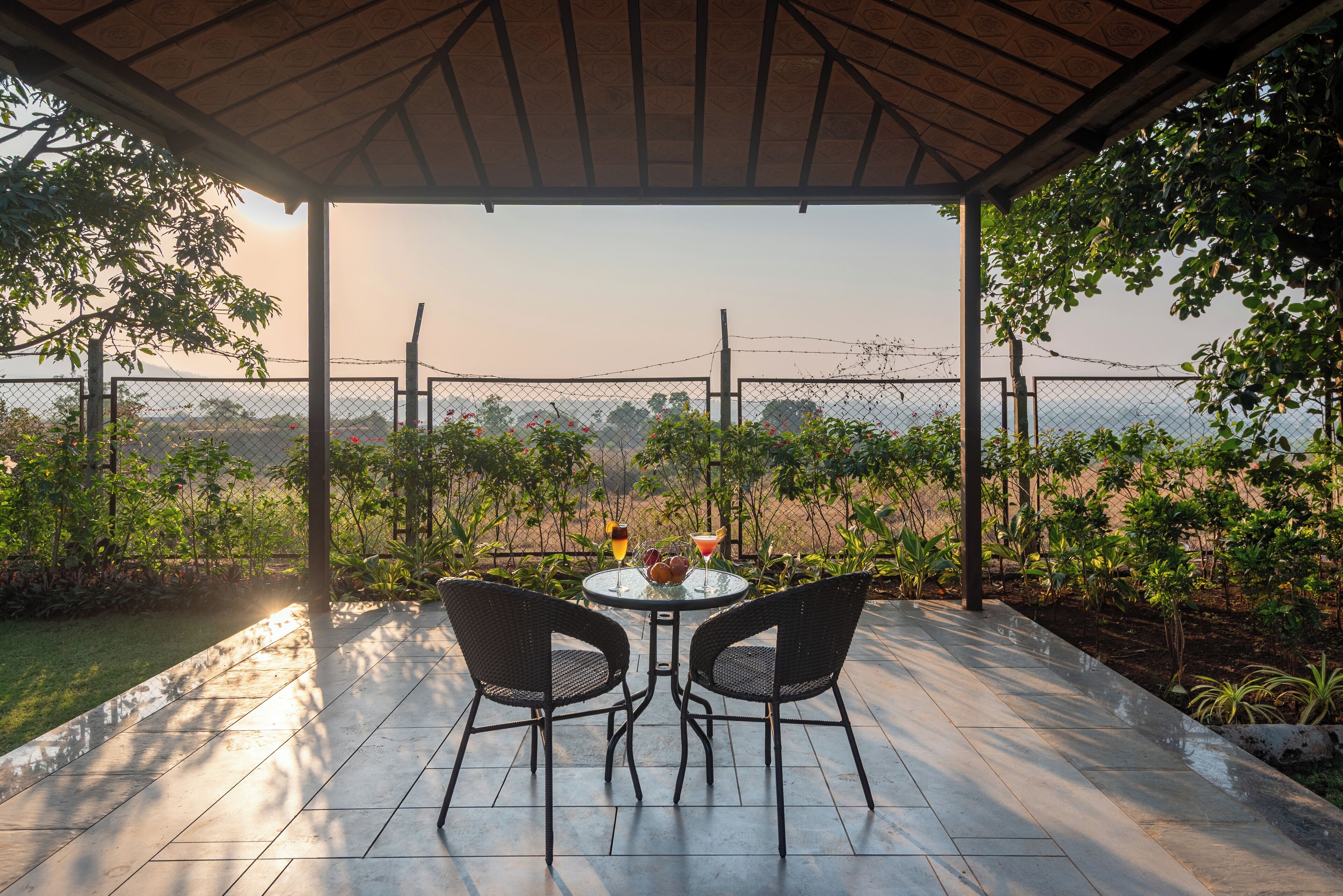 A covered patio with two chairs and a table, overlooking a fenced area with plants and a distant view.