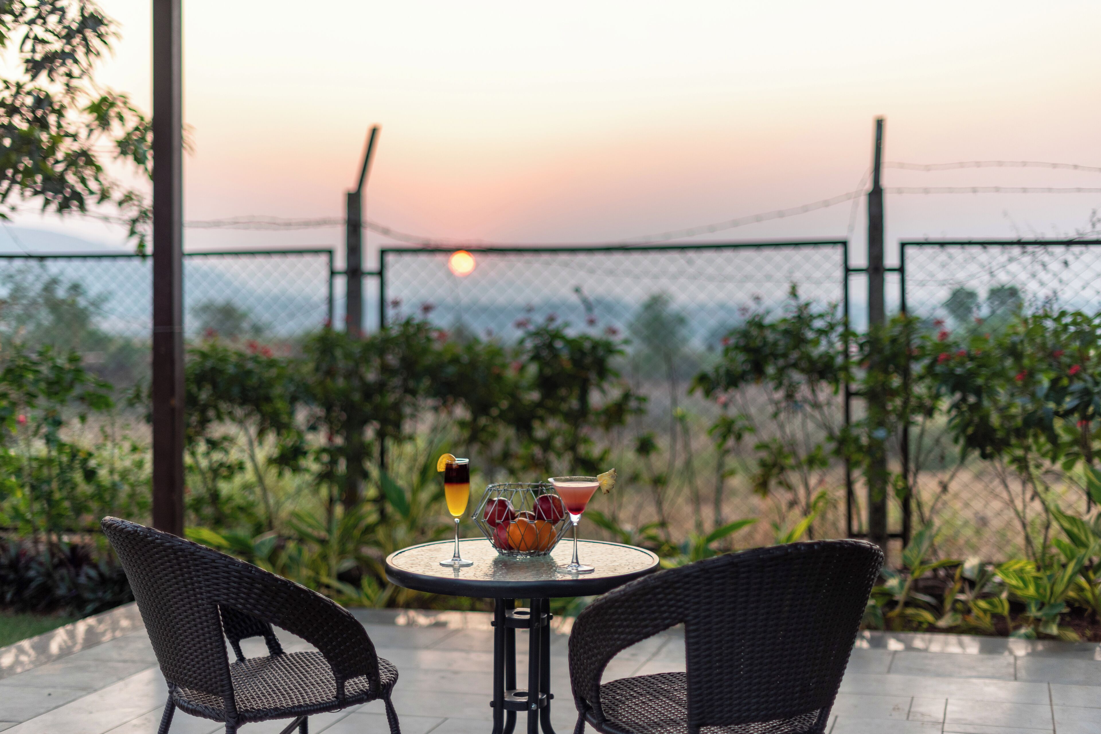 Una mesa redonda con dos sillas de mimbre, un cuenco de fruta y dos bebidas en un patio con vista al atardecer y una valla de alambre en el fondo.