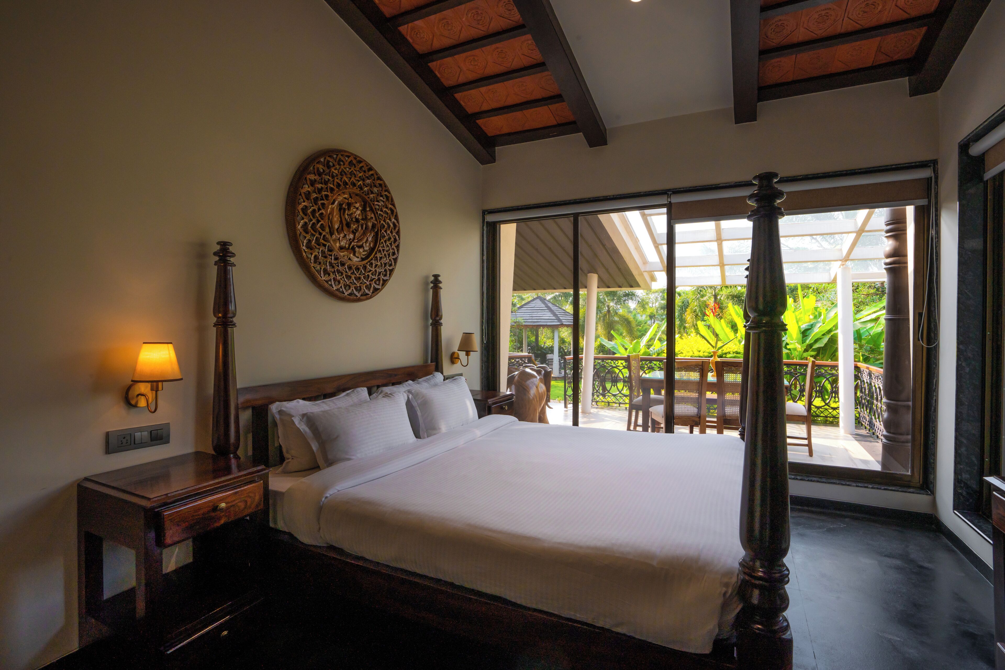 A spacious bedroom with a large bed, a nightstand with a lamp, a decorative wall piece, and a view of a lush garden through a sliding glass door.