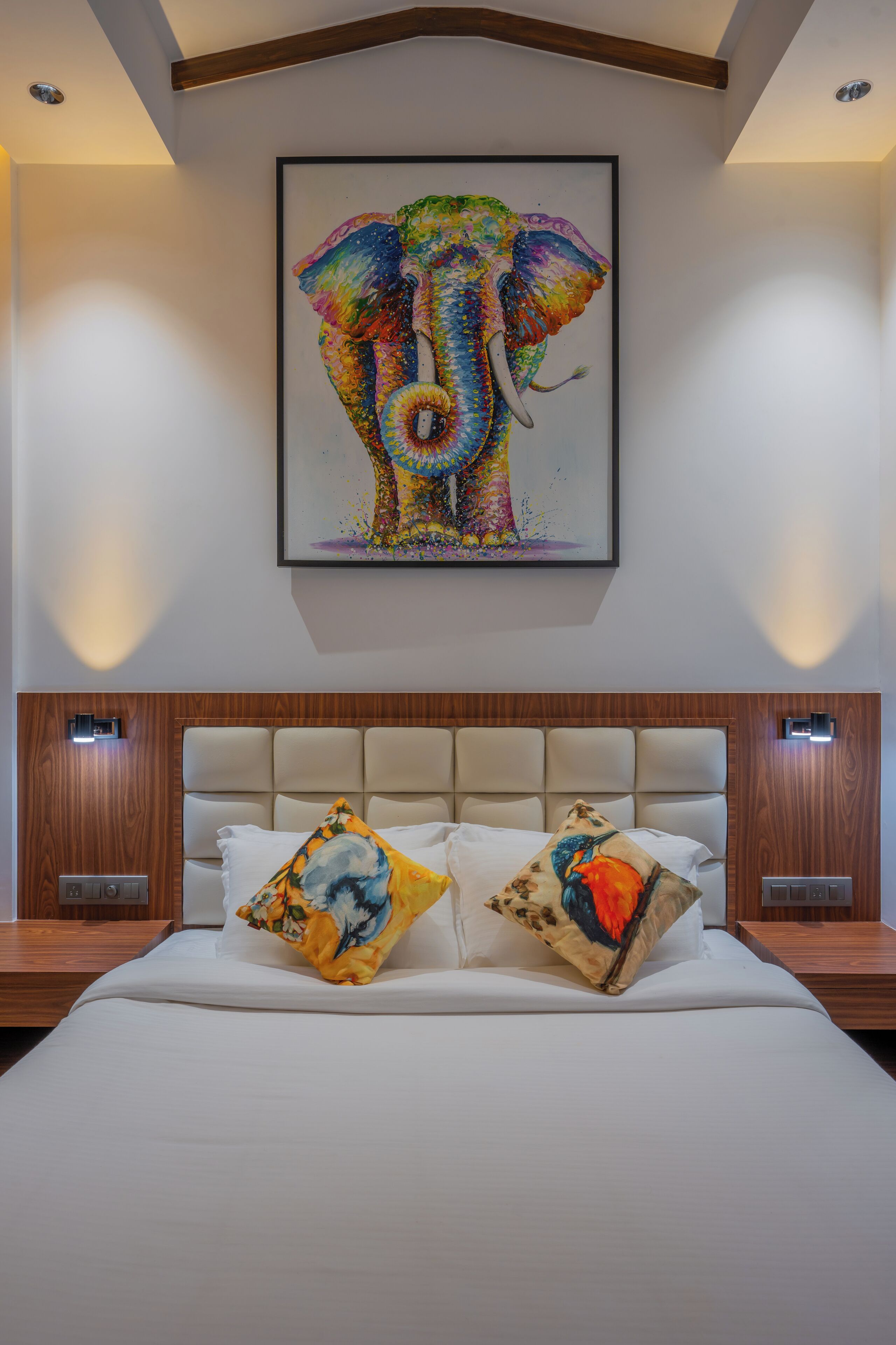 A bedroom with a large bed, two pillows with colorful artwork, a wooden headboard, and a framed elephant painting on the wall.