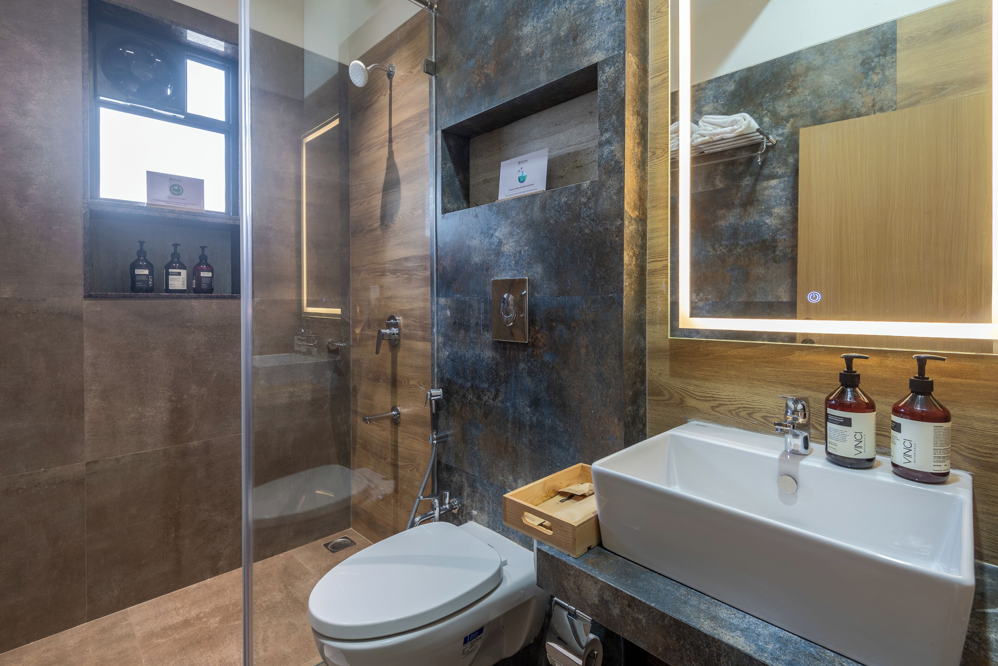A modern bathroom with a glass shower enclosure, a white toilet, a sink with two bottles of hand soap, and a wooden box on the countertop.
