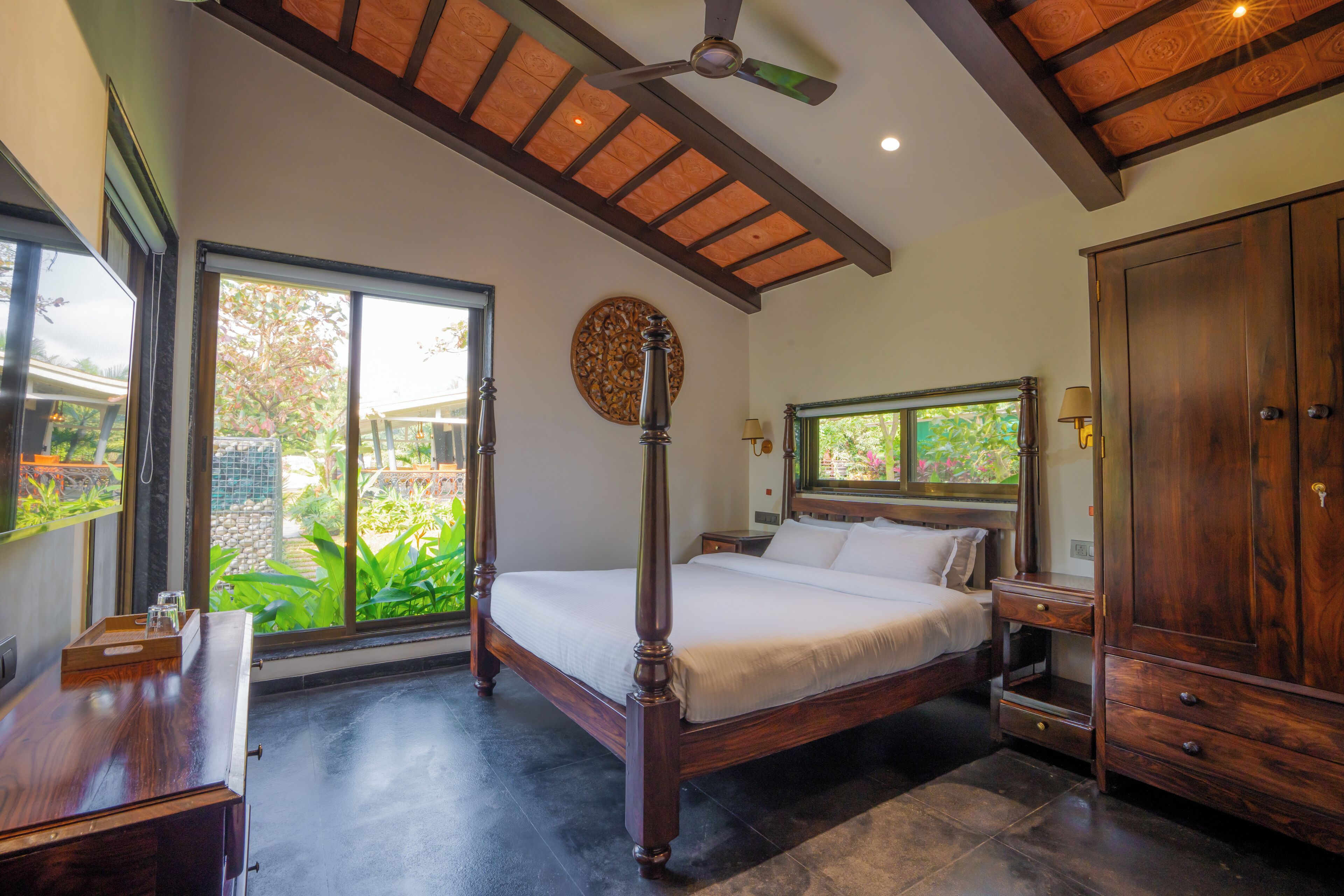 A bedroom with a large bed, wooden furniture, a ceiling fan, and a view of greenery outside.
