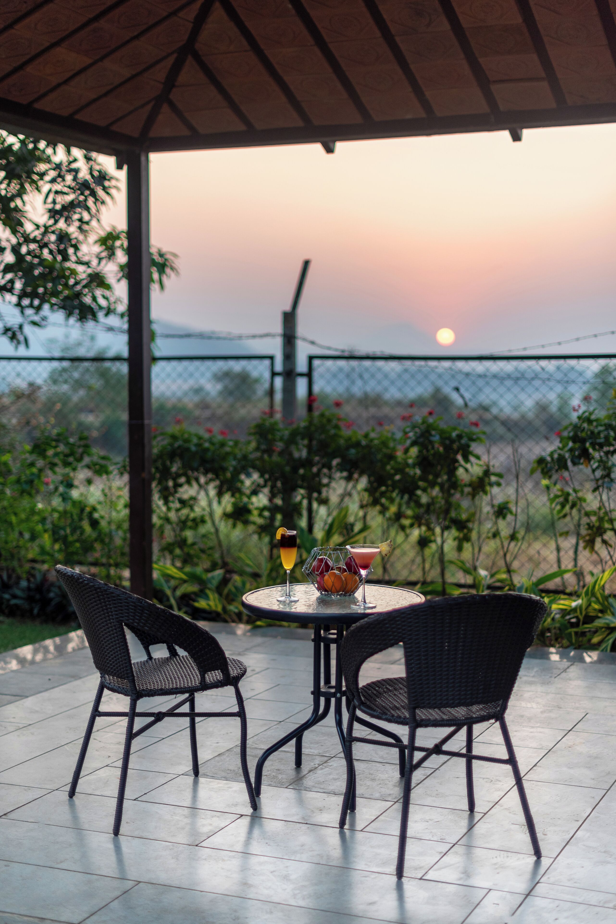 A covered patio with two wicker chairs and a table, a sunset view, and a fence with greenery.