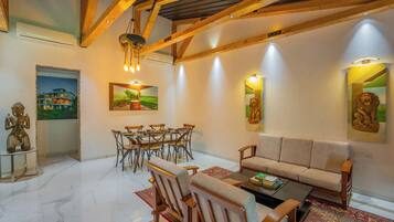 A spacious living room with a high ceiling, wooden beams, a sofa, a coffee table, a dining area with chairs, and artwork on the walls.