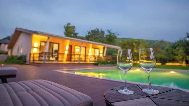 Un área junto a la piscina con dos copas de vino vacías sobre una mesa de madera, una silla de descanso y un edificio con ventanas iluminadas al fondo.