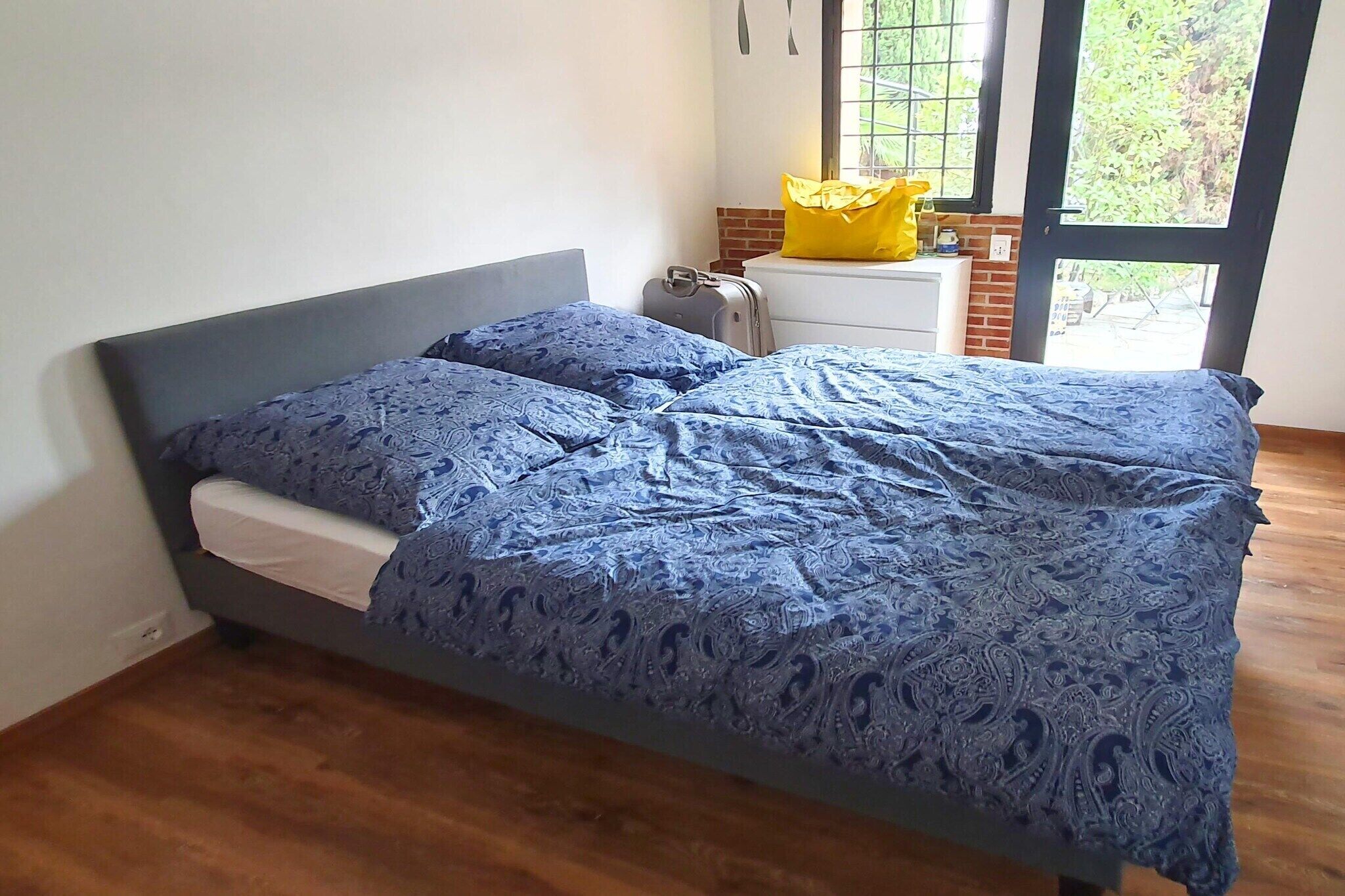 A bed with a patterned bedspread, a suitcase, a yellow pillow, and a window with a view of greenery.