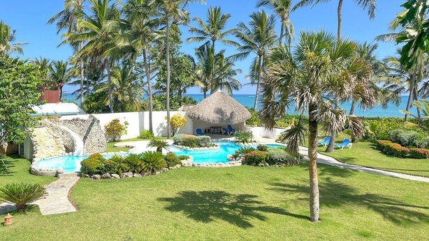 A tropical resort with a swimming pool, palm trees, and a thatched-roof hut. - All-inclusive 3bd beachfront central condo with sea views! (Cabarete)