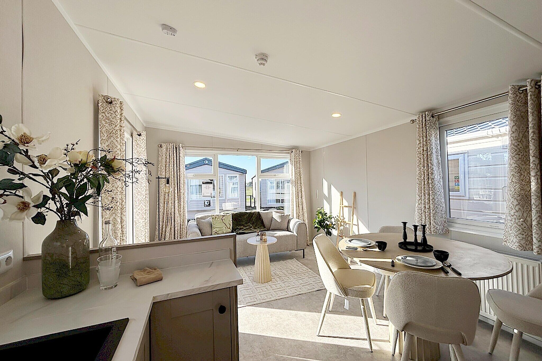 Ein modernes Wohnzimmer mit Sofa, Couchtisch und Essbereich. Auf der Anrichte steht eine Vase mit Blumen, und es gibt ein Fenster mit Vorhängen.
