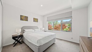 Een moderne slaapkamer met een groot bed, nachtkastjes, een raam met uitzicht op herfstbomen en twee kunstwerken aan de muur.