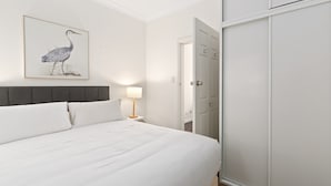 Een netjes opgemaakt bed met witte lakens, een grijze hoofdbord en een lijstwerk met een afbeelding van een vogel aan de muur.