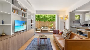 Una sala moderna con televisor, sofá y mesa de centro. Se ve una estantería, un comedor y una cocina.