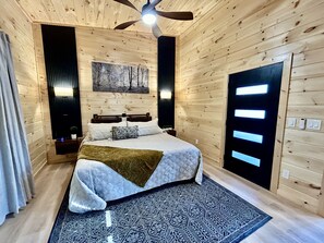 A bedroom with a bed, a ceiling fan, a painting, and a door with illuminated panels. - BRAND NEW Mid-Century Modern Cabin with Hot Tub, Fire Pit & Outdoor Fireplace (Sevierville)