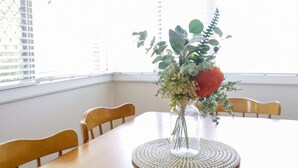 A dining table with a clear vase of flowers, wooden chairs, and a window with blinds.