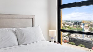Una camera d'albergo con un letto, un comodino, una lampada e un'ampia finestra con vista sulla città.