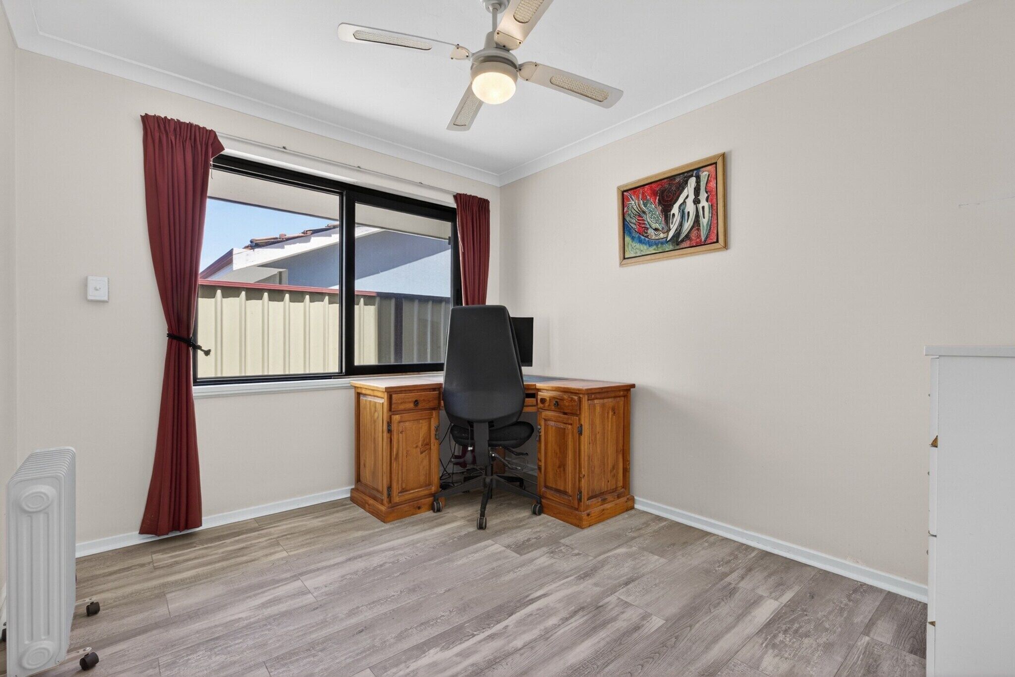 A room with a wooden desk, a black office chair, a ceiling fan, a window with red curtains, and a colorful painting on the wall.