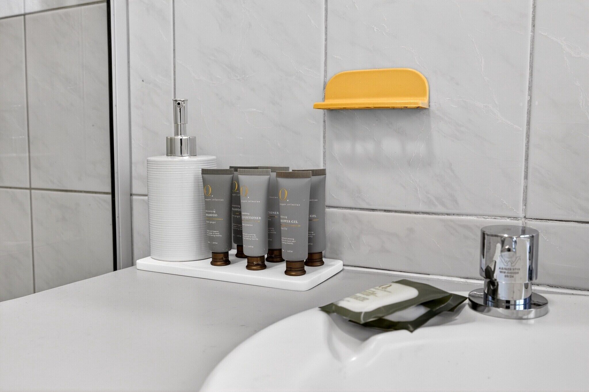 Une salle de bain avec un lavabo blanc, un porte-savon jaune et trois flacons gris d’après-shampoing disposés sur un plateau blanc.