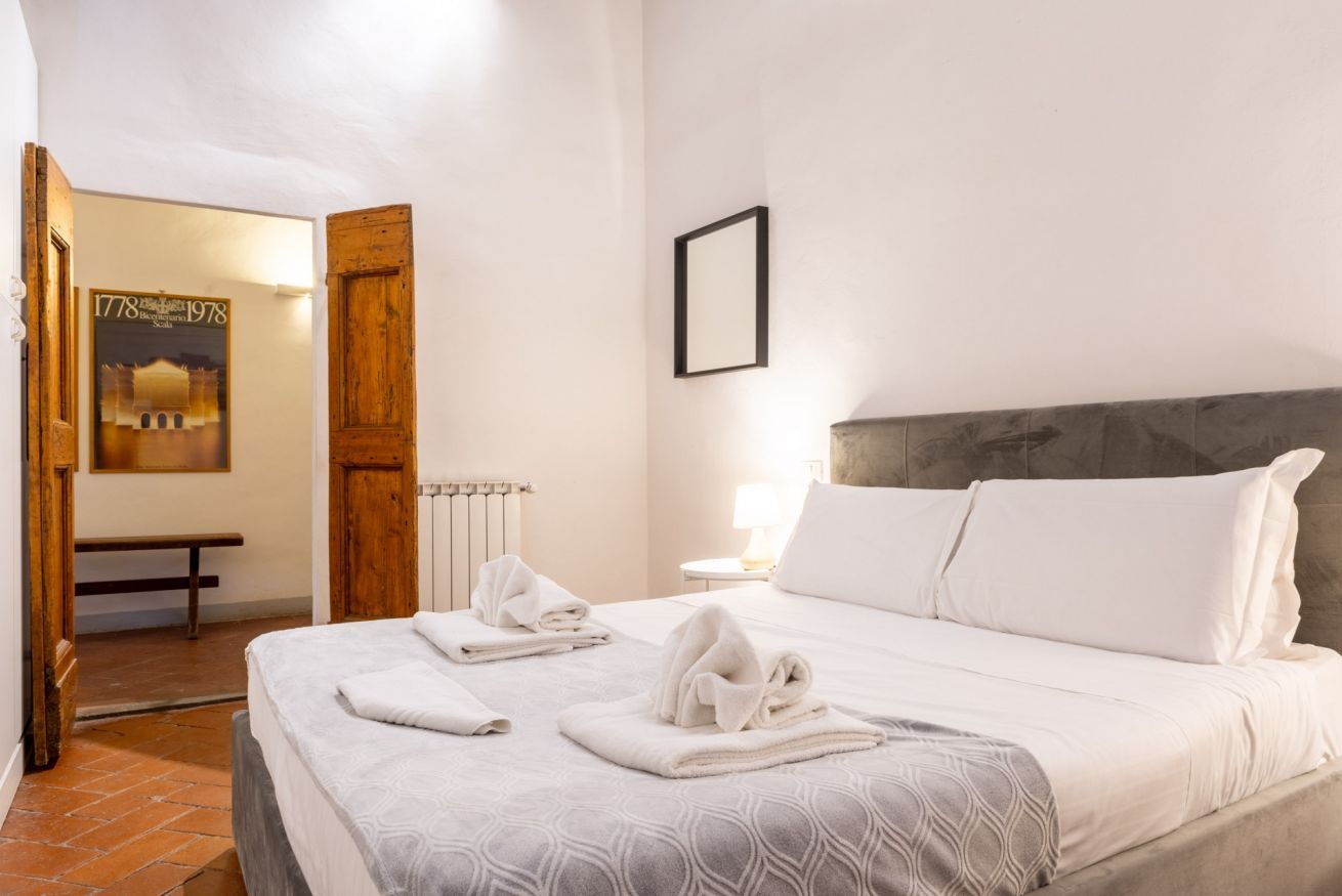 Una cama bien hecha con ropa de cama y toallas blancas, una puerta de madera, un banco y un cuadro en la pared.