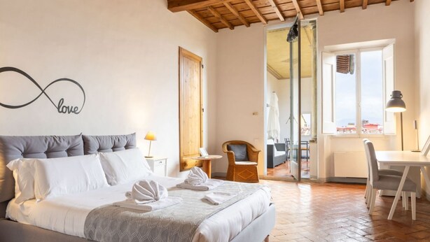 Un dormitorio con cama, una pared decorada con el símbolo infinito 'love', una zona de comedor con mesa y sillas, y vistas a través de un ventanal.