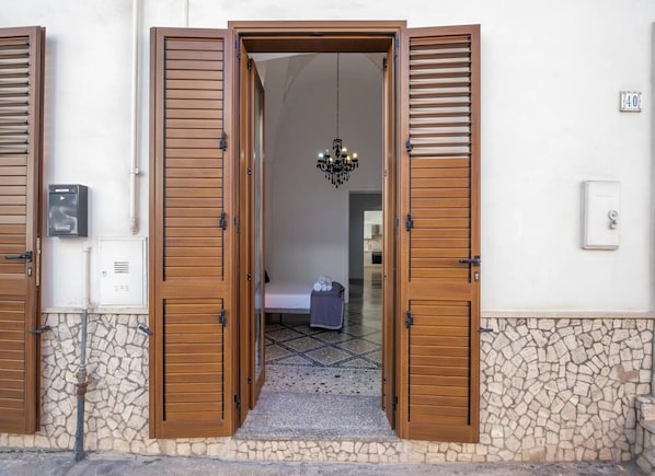Uma porta com portões de madeira, parede de pedra, chão de azulejos, cama com coberta e um candelabro.
