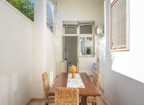 A narrow corridor with a wooden table and wicker chairs, a door leading to a room, and a window with a plant.