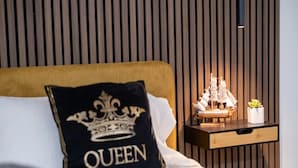 Un lit avec un oreiller portant l’inscription « QUEEN », une table de chevet ornée d’une maquette de bateau et un mur recouvert de panneaux de bois verticaux.