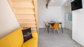 Un intérieur moderne avec un escalier en bois, un canapé jaune agrémenté d’un coussin, une table à manger avec des chaises et un coin cuisine.
